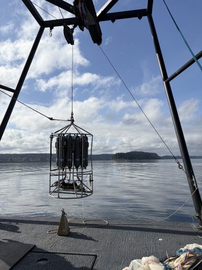 A metal oceanographic instrument is suspended above calm waters on a clear day. A-frame structure holds the device; distant islands are visible.