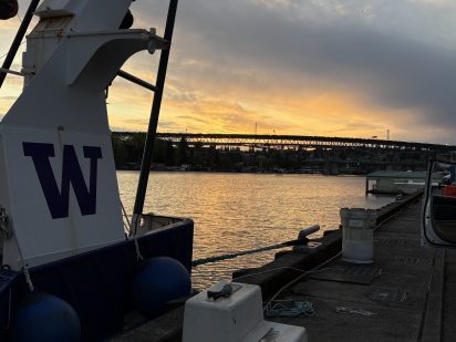 Sunset over a calm river with a large "W" on a boat in the foreground. A bridge spans the background under a sky with warm orange hues. Serene atmosphere.
