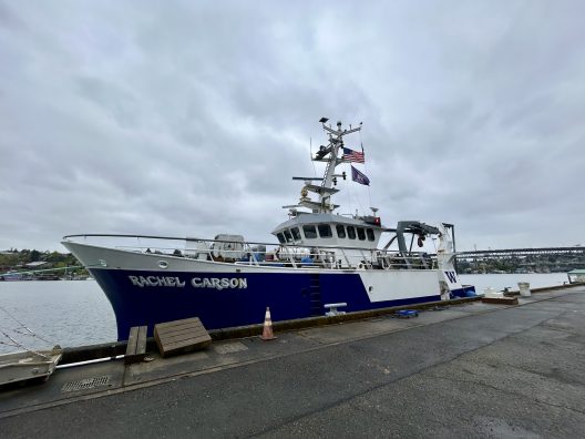 A large blue and white research vessel named "Rachel Carson" is docked at a pier. The scene is overcast, and the American flag atop the ship is visible.