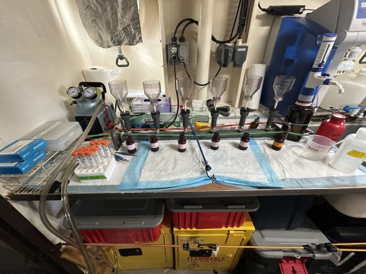 A laboratory bench with a setup for filtration. Multiple funnels and flasks are arranged in a line, with tubes connecting them. Nearby, there are various lab materials including test tubes, protective gloves, and bottles. The organization suggests meticulous scientific work.