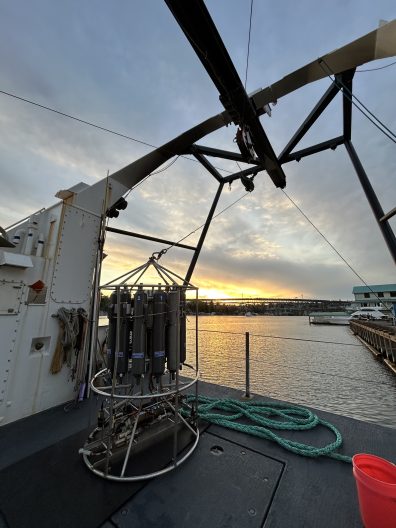 Sunset view from a ship's deck with scientific equipment and coiled ropes. The sky is vibrant with warm hues, casting reflections on the water.