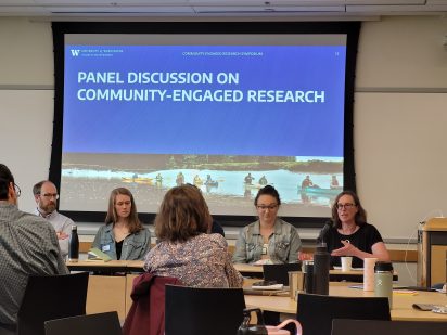 A panel of five people discusses community-engaged research in a conference room with a large screen displaying the topic and an image of kayakers.