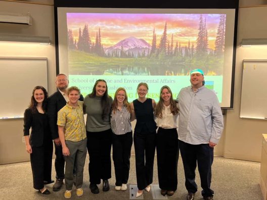 A group of eight smiling people stands in front of a classroom screen displaying a mountain and forest landscape. The atmosphere is cheerful and professional.