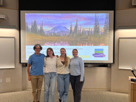 Four people standing in front of a presentation screen displaying a scenic landscape. The presentation is for the School of Marine and Environmental Affairs at the University of Washington. The group appears happy and relaxed.