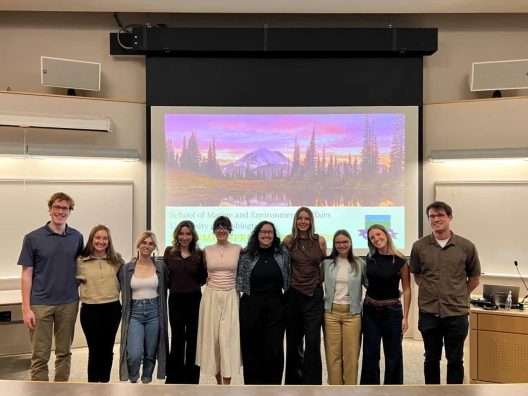 A group of nine smiling people stands in front of a screen displaying colorful landscape art. They're in a lecture hall, exuding a cheerful and collaborative atmosphere.