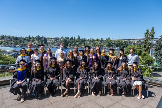 Graduates in black gowns and caps smile in a group photo outdoors, with a scenic background of water, trees, and a clear blue sky. Celebratory mood.