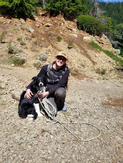 A person kneels on rocky terrain, smiling beside a black and white dog. The background features a hillside with green trees and shrubs, under a clear blue sky.