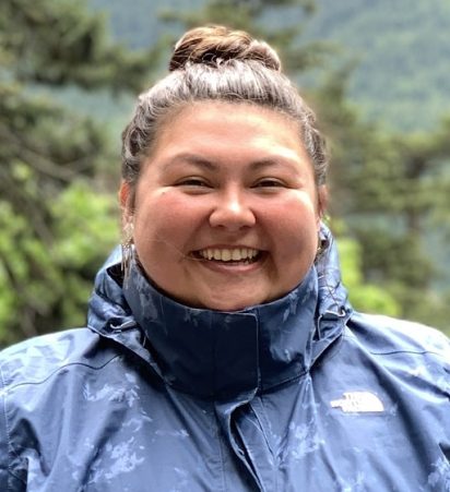 Smiling woman in a blue jacket with a forested, mountainous background. She appears content and joyful, conveying a sense of outdoor adventure.
