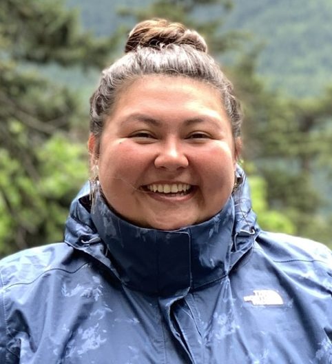 Smiling woman in a blue jacket with a forested, mountainous background. She appears content and joyful, conveying a sense of outdoor adventure.