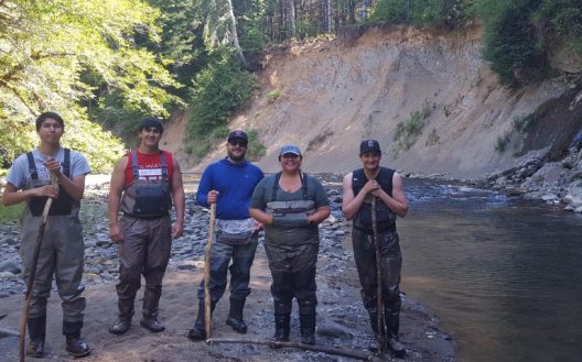 Five people stand smiling by a calm river, wearing waders and holding walking sticks, surrounded by lush trees and a steep, earthy riverbank.