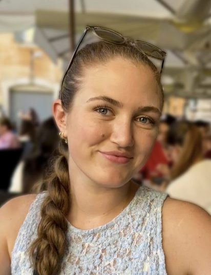 Smiling woman with braided hair and glasses on head, wearing a light blue lace top. Background shows people seated outdoors, creating a cheerful vibe.