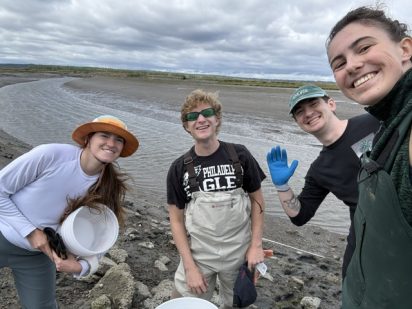Four smiling friends pose on a muddy coastal shoreline under cloudy skies, wearing outdoor gear; one waves in a bright blue glove.