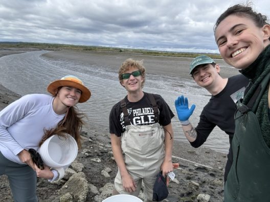 Four smiling friends pose on a muddy coastal shoreline under cloudy skies, wearing outdoor gear; one waves in a bright blue glove.
