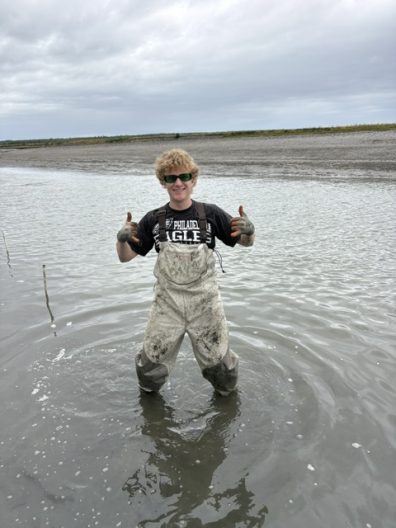 Smiling person in muddy chest waders gives two thumbs up while standing in shallow tidal water under a cloudy sky.