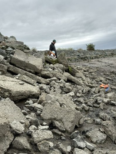 Hiker in dark clothing traverses a rugged gray volcanic rock field beneath overcast skies; bright orange gear rests among stones nearby.