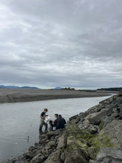 Three people gather on a rocky tidal shoreline beside a calm channel beneath a vast overcast sky, creating a quiet, reflective mood.