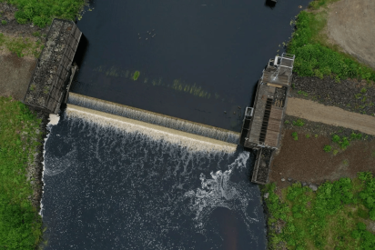 Aerial view of a small concrete dam spilling into a dark river, with stone banks, green vegetation, dirt paths, and foaming white water.