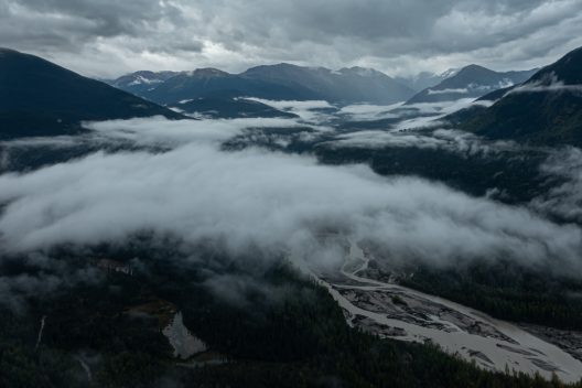 Aerial view of a mountain valley blanketed in low clouds, with a winding river below and dark peaks under a moody, overcast sky.