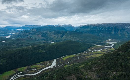 Aerial view of a braided river winding through a vast forested mountain valley beneath a dramatic, cloudy sky, creating a remote, tranquil mood.