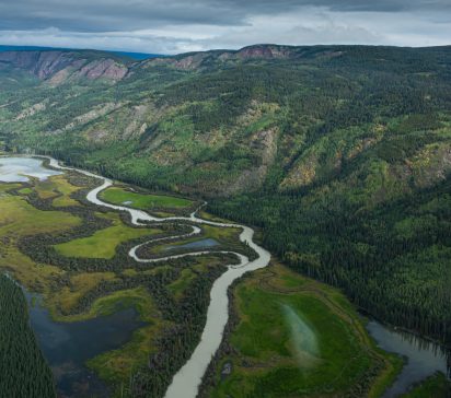 A winding river snakes through lush green wetlands beneath forested mountain slopes and a cloudy sky, creating a vast, tranquil wilderness scene.