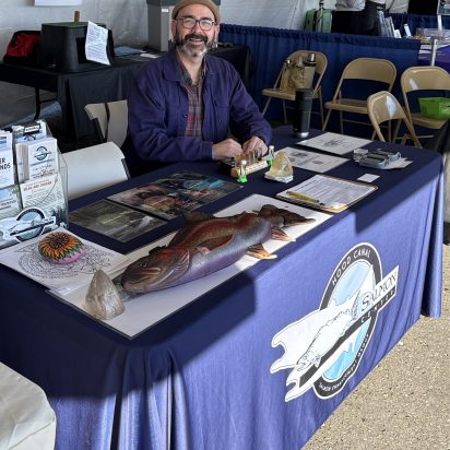 Smiling naturalist at a Hood Canal salmon display, seated behind a blue-covered table with a lifelike fish model, fossils, and educational materials.